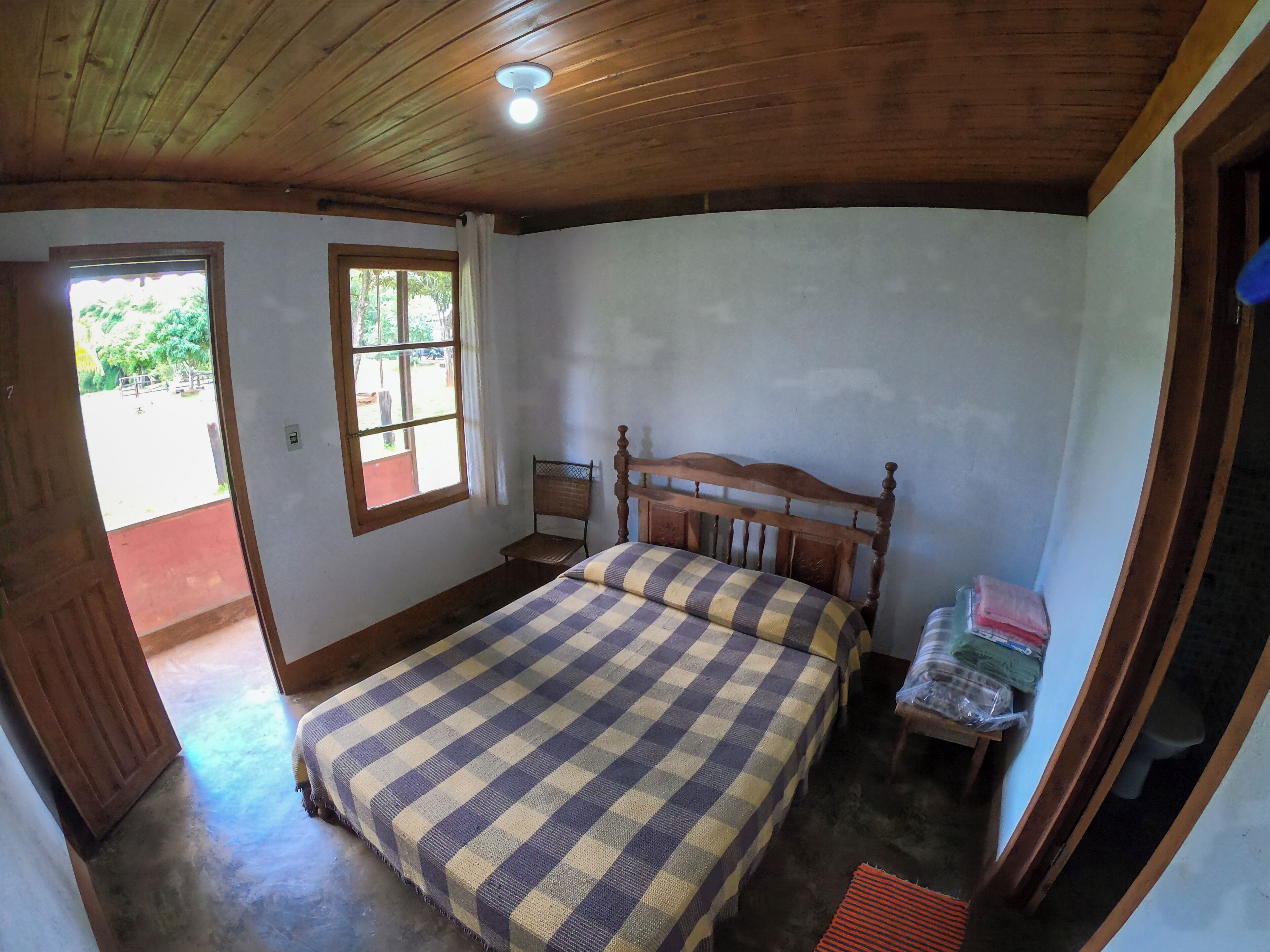 Une chambre avec un plafond en bois, un lit recouvert d’une couverture à carreaux, une fenêtre avec des rideaux et une porte donnant sur l’extérieur.
