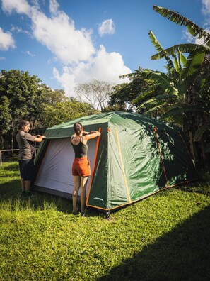 Uma tenda verde com detalhes em laranja em uma área gramada, com árvores e céu claro.