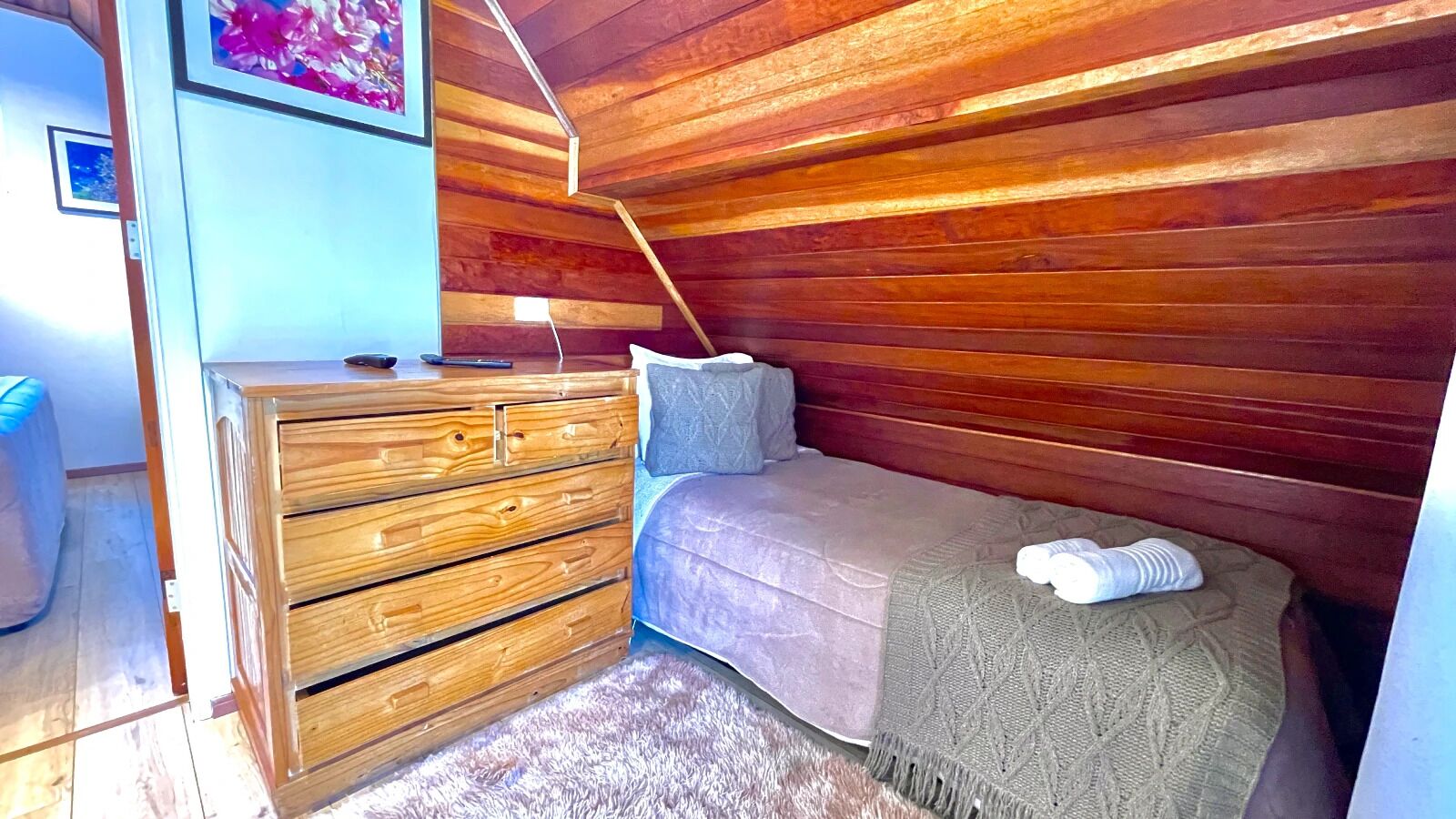 A bedroom with a wooden headboard, a bed with a grey blanket, a wooden dresser, and a small bedside table with a lamp.