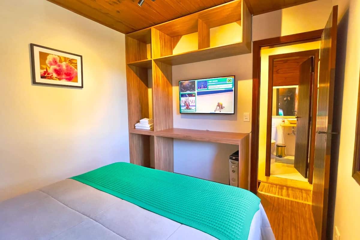 A hotel room with a bed, a TV mounted on a wooden shelf, a green mat, and a framed picture on the wall.