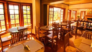 A dining area with multiple wooden tables and chairs, large windows with views of greenery, and a fireplace. - Hortelã Village (Campos do Jordão)