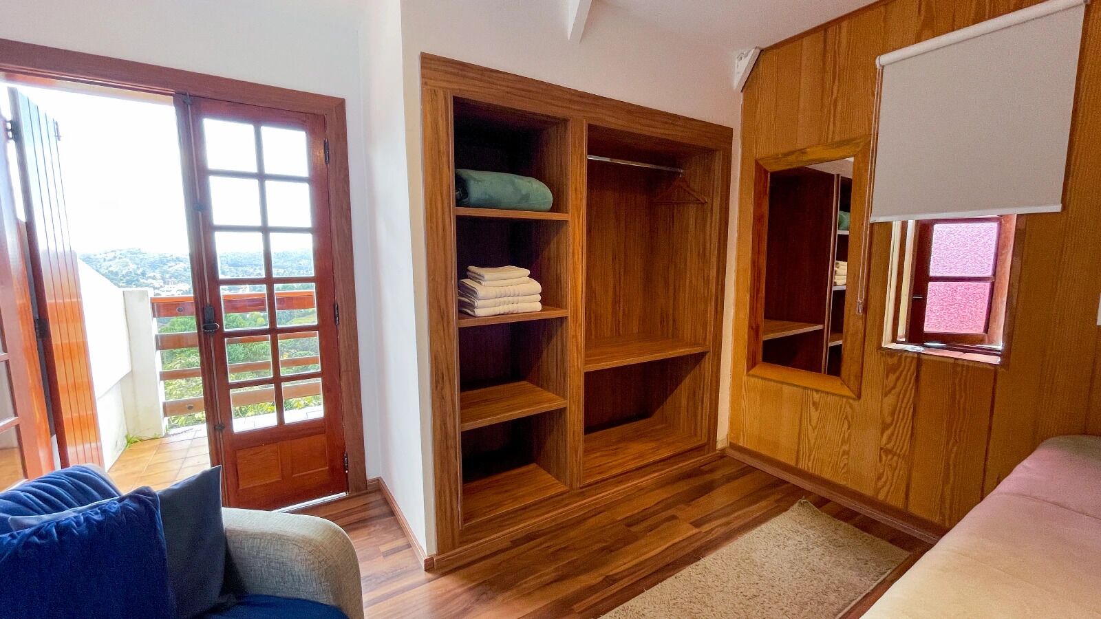 A room with a wooden wardrobe, a sliding glass door leading to a balcony, and a bed with a white mattress.