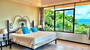 A bedroom with a bed, a nightstand, a lamp, and a window with a view of greenery. - Villa Grande – Spacious 5-Bedroom Retreat with Private Pool in Manuel Antonio (Quepos)