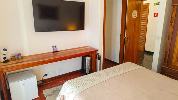 A hotel room with a bed, a desk with a computer, a TV mounted on the wall, and a small refrigerator. - Pousada Hortelã (Campos do Jordão)