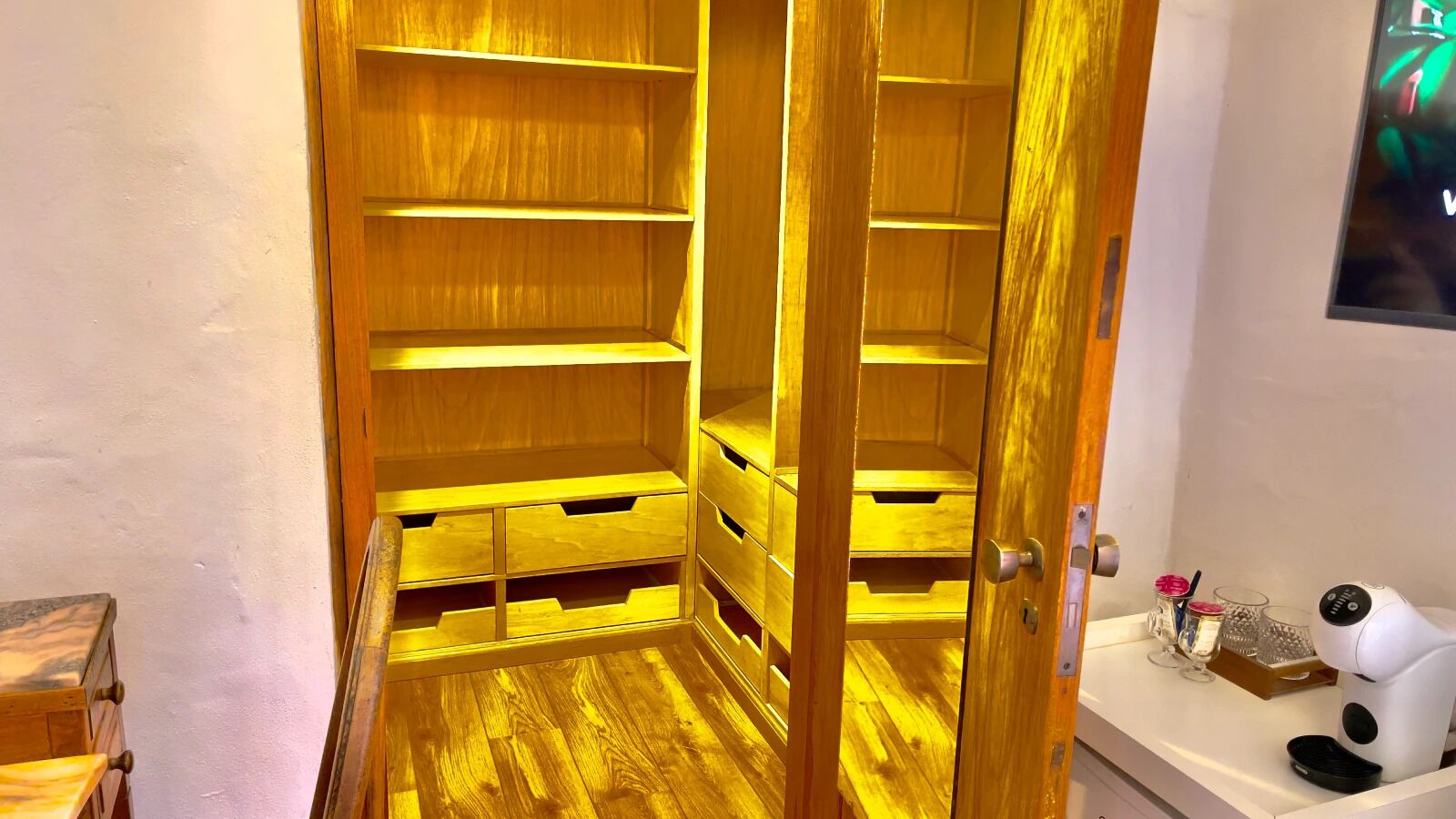 Une armoire en bois avec plusieurs étagères et tiroirs, un mur blanc et une petite table sur laquelle se trouvent un sèche-cheveux et des pots.