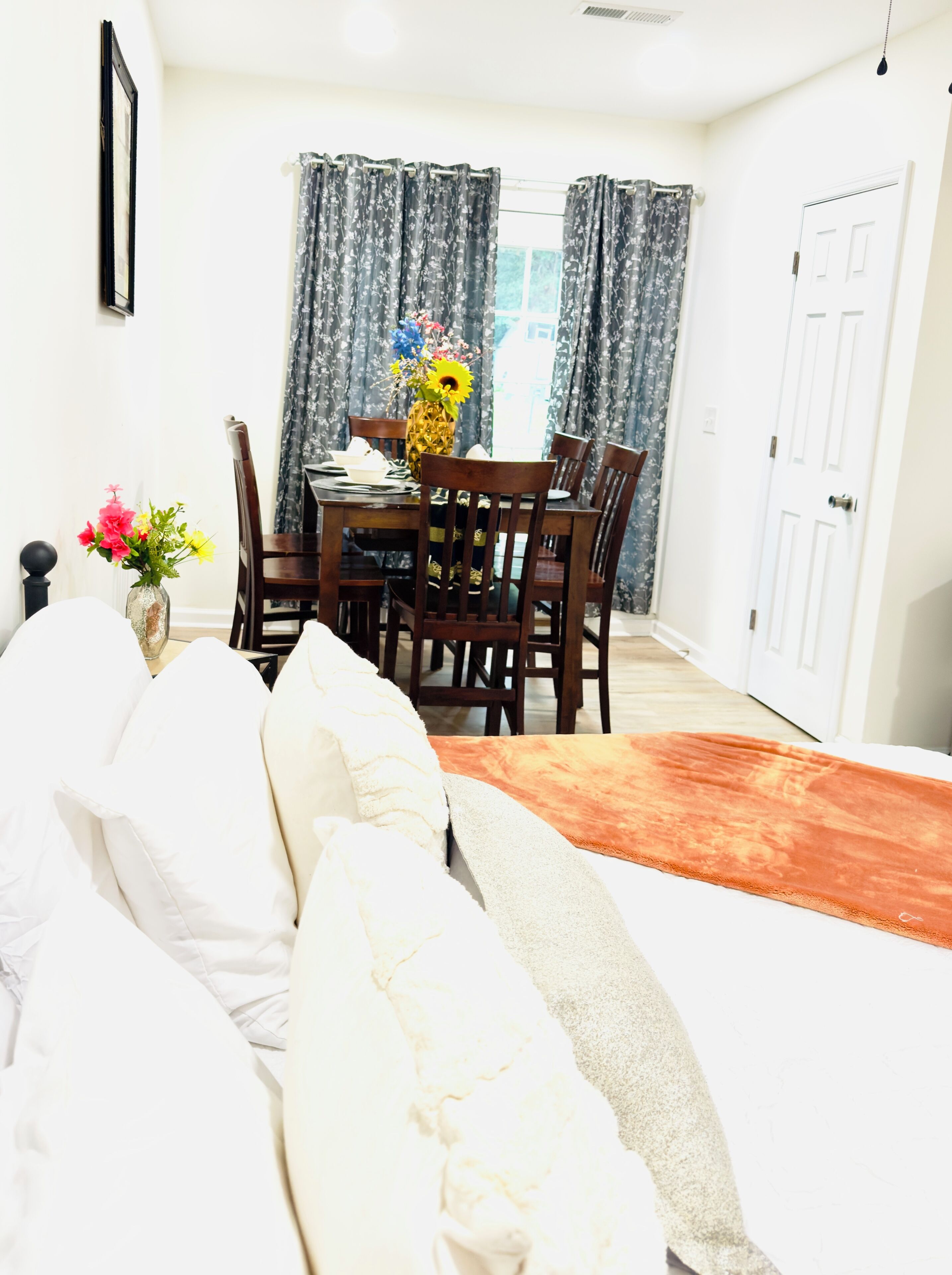 Une chambre à coucher avec un lit, une table à manger avec des chaises, un vase avec des fleurs et une porte.