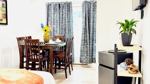 A hotel room with a bed, a dining table with chairs, a ceiling fan, a TV mounted on the wall, and a potted plant.