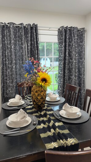A dining table set with white plates, a vase with flowers, and a view of a house through a window with curtains. - Modern & Cozy Summerville Apt - Fully Equipped (Summerville)