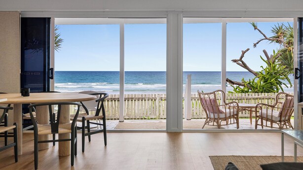 Une pièce dotée d’une grande porte-fenêtre coulissante donnant sur un balcon avec vue sur l’océan. À l’intérieur, on trouve une table en bois avec des chaises et un petit coin cuisine.