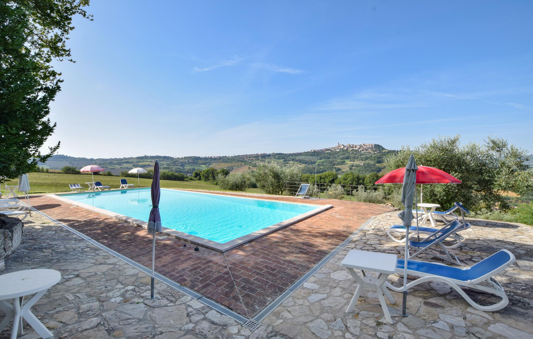 A rectangular swimming pool with a brick border, surrounded by lounge chairs and umbrellas, set against a backdrop of hills and a clear sky.