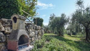 A stone-built outdoor oven with a chimney, surrounded by olive trees and greenery.