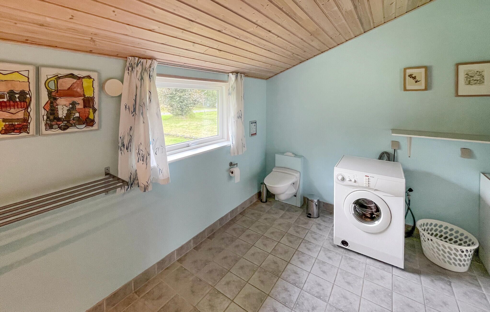 A laundry room with a washing machine, a toilet, and a window with curtains.
