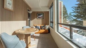 A cozy room with a wooden wall, a round wooden table, a blue chair, a sofa with cushions, and a large window with a view of trees and snow. - Canyon View at Canyon Lodge (Mammoth Lakes)