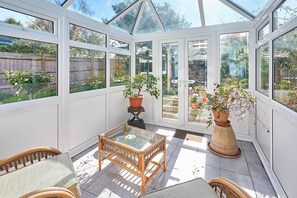 A conservatory with white-framed glass doors, wicker furniture, and potted plants. - Host Stay Bodiam Close (Seaford)
