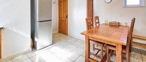 A kitchen with a stainless steel refrigerator, a wooden dining table with chairs, a clock on the wall, and a window providing natural light.