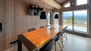 A spacious dining area with a long wooden table, black chairs, and pendant lights. A TV is mounted on the wall, and large windows offer a view of the outdoors. - Lovely home in Gålå with sauna (Gålå)