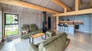 A modern living room with a sofa, ottoman, and a coffee table. There is a fireplace, a kitchen area with bar stools, and a large window with a view of greenery. - Lovely home in Gålå with sauna (Gålå)