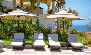 Un espace aménagé au bord de la piscine, avec des chaises longues et des parasols, entouré d’une végétation luxuriante et offrant une vue sur l’océan.