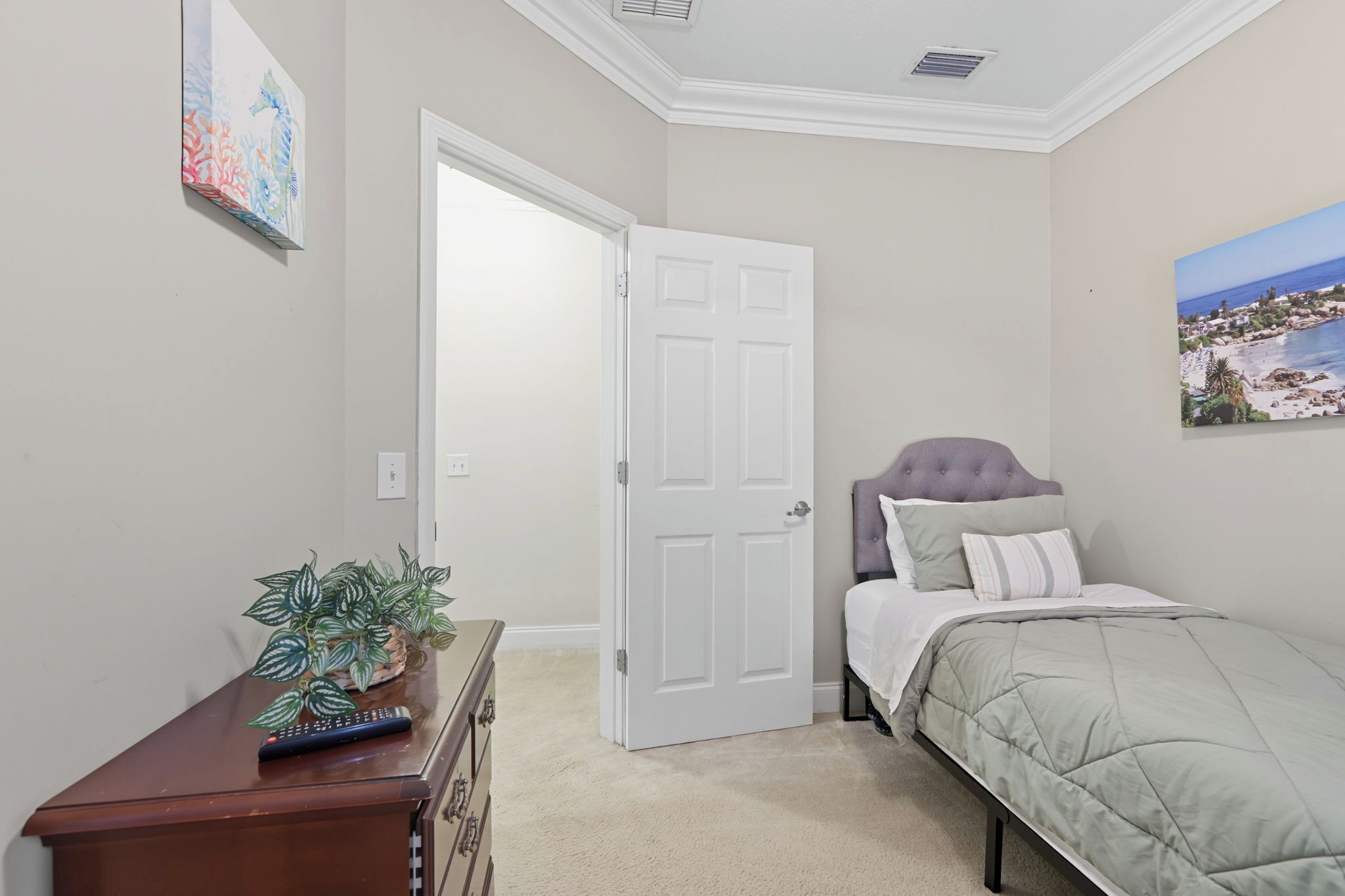 Une chambre à coucher avec un lit, une commode ornée d’une plante, une porte et un tableau accroché au mur.