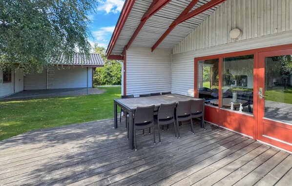 En udendørs terrasse med et træbord og stole, en glasdør der fører til en patio, og en bygning i baggrunden.