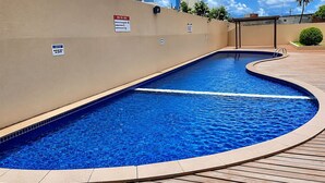 A swimming pool with a curved edge, surrounded by a wooden deck and beige walls. - Hosts BR - Flat frente mar (Fortaleza)