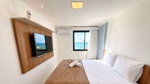 A hotel room with a bed, a TV mounted on the wall, a window with a view of the sea, and an air conditioning unit. - Hosts BR - Flat frente mar (Fortaleza)