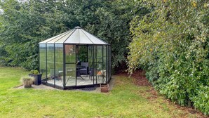 A glass conservatory with a hexagonal shape, a small table, and two chairs inside, surrounded by a grassy yard and dense foliage. - Pet friendly home in Sønder Felding with WiFi (Sønder Felding)