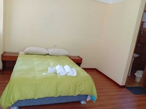 Una cama individual con una colcha verde, dos almohadas, una mesita de noche de madera y un par de toallas enrolladas.