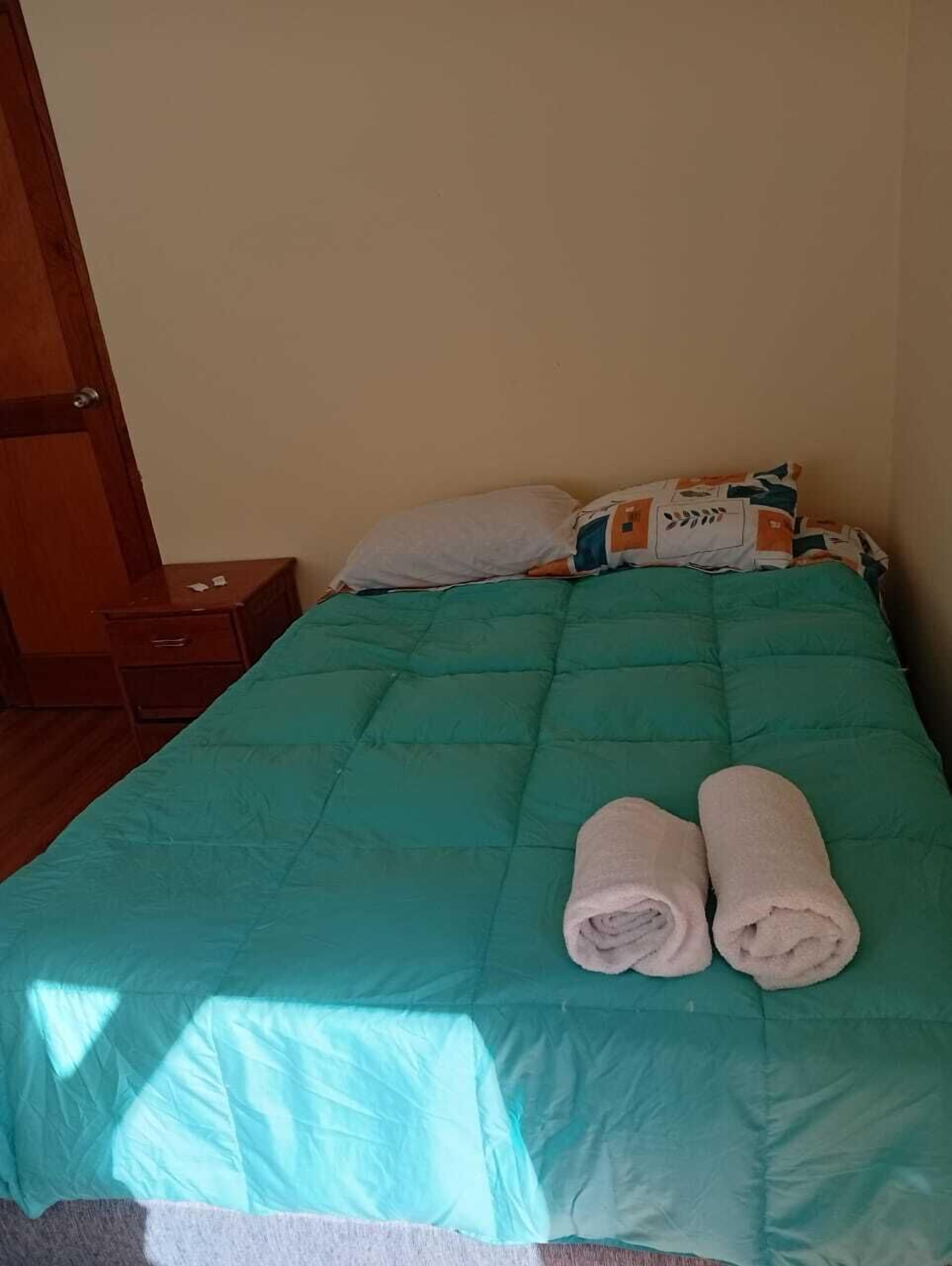 A bed with a teal bedspread, two rolled towels, and a pillow with a leaf pattern.