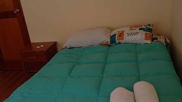 A bed with a teal bedspread, two rolled towels, and a pillow with a leaf pattern.