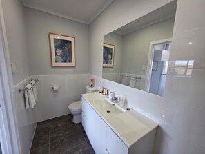 Uma casa de banho moderna com uma pia branca, sanita e um espelho grande. Existem duas obras de arte emolduradas na parede, e um porta-toalhas é visível.