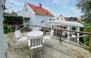 Una terraza de madera con sillas Adirondack blancas y una mesa redonda, con vistas a una zona residencial con casas y vegetación.