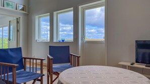 A room with a bed, two chairs, a TV, and large windows with a view of the sky and clouds.
