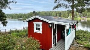 A small red cabin with a white door and window, situated by a lake with a wooden dock extending into the water. - Beautiful home in Säffle with lake view (Säffle)