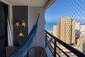 A balcony with a hammock, a chair, and a view of buildings through a chain-link fence.