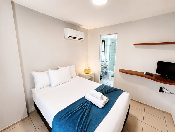 A hotel room with a bed, a TV on a wooden shelf, and an air conditioning unit on the wall.