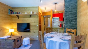 A cozy wooden room with a television, a dining table set for four, and a kitchen area with red cabinets. - RESIDENCE ORIONDE - L'OURSON (LES DESERTS)
