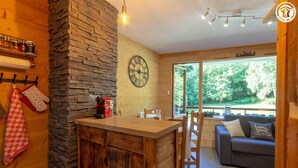 A cozy room with a stone wall, wooden furniture, a clock, and a large window offering a view of greenery. - RESIDENCE ORIONDE - L'OURSON (LES DESERTS)