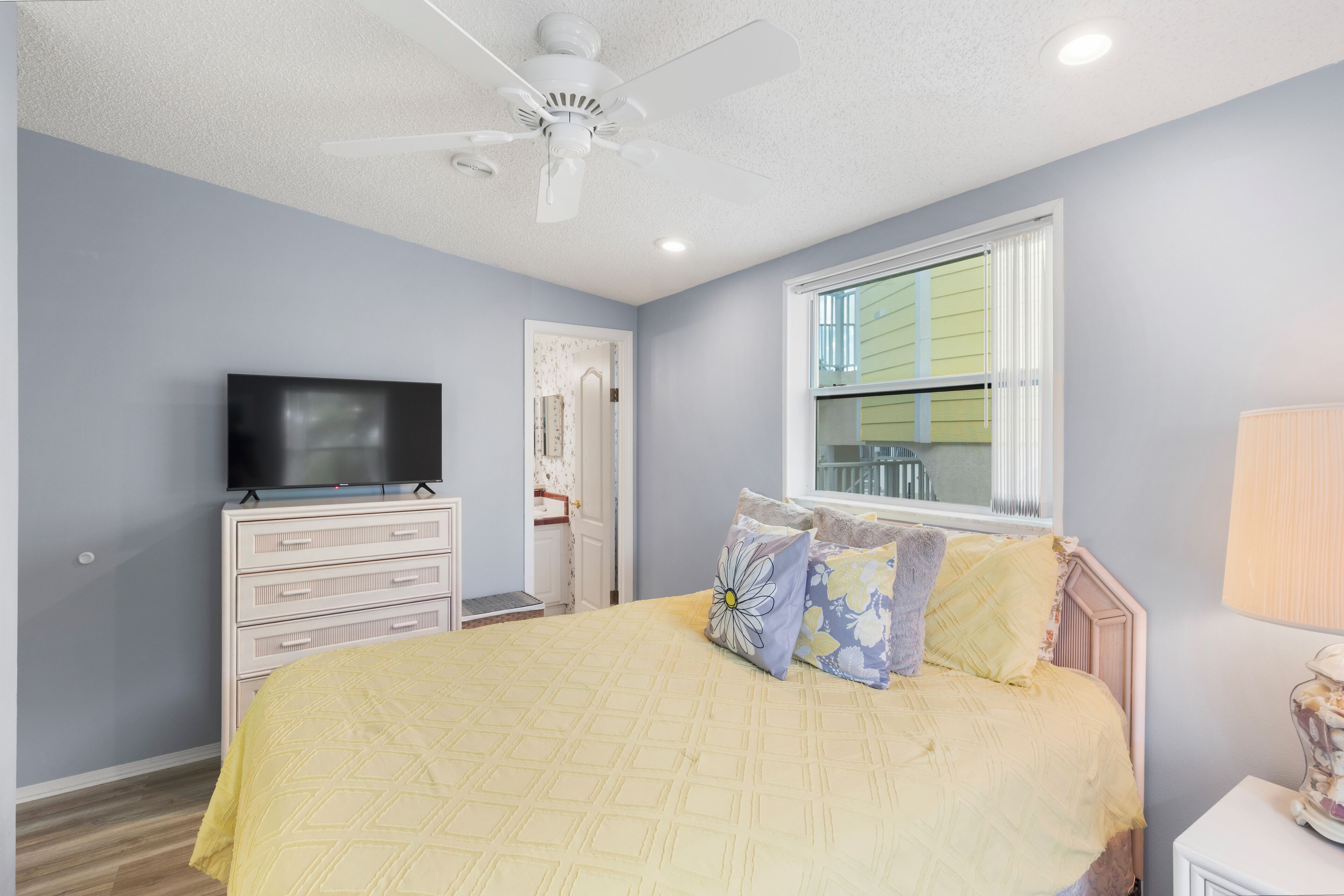 A bedroom with a bed, a television on a dresser, a ceiling fan, and a window with blinds.