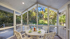 An outdoor dining area with a round table and white chairs, surrounded by glass walls with a view of a pool and greenery. - KIAWAH, West Beach, 5BR Luxury Home, POOL & Incredible PORCHES! (Kiawah Island)