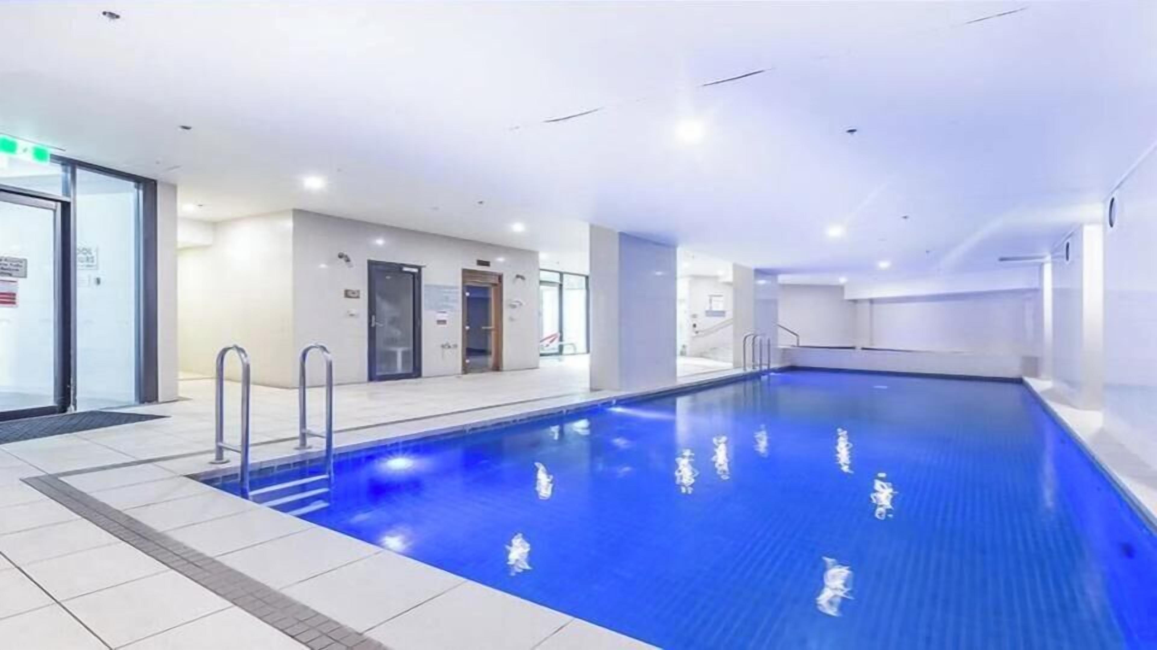 En stor indendørs swimmingpool med klart, blåt vand, omgivet af flisegulv og vægge, samt metalgelændere.