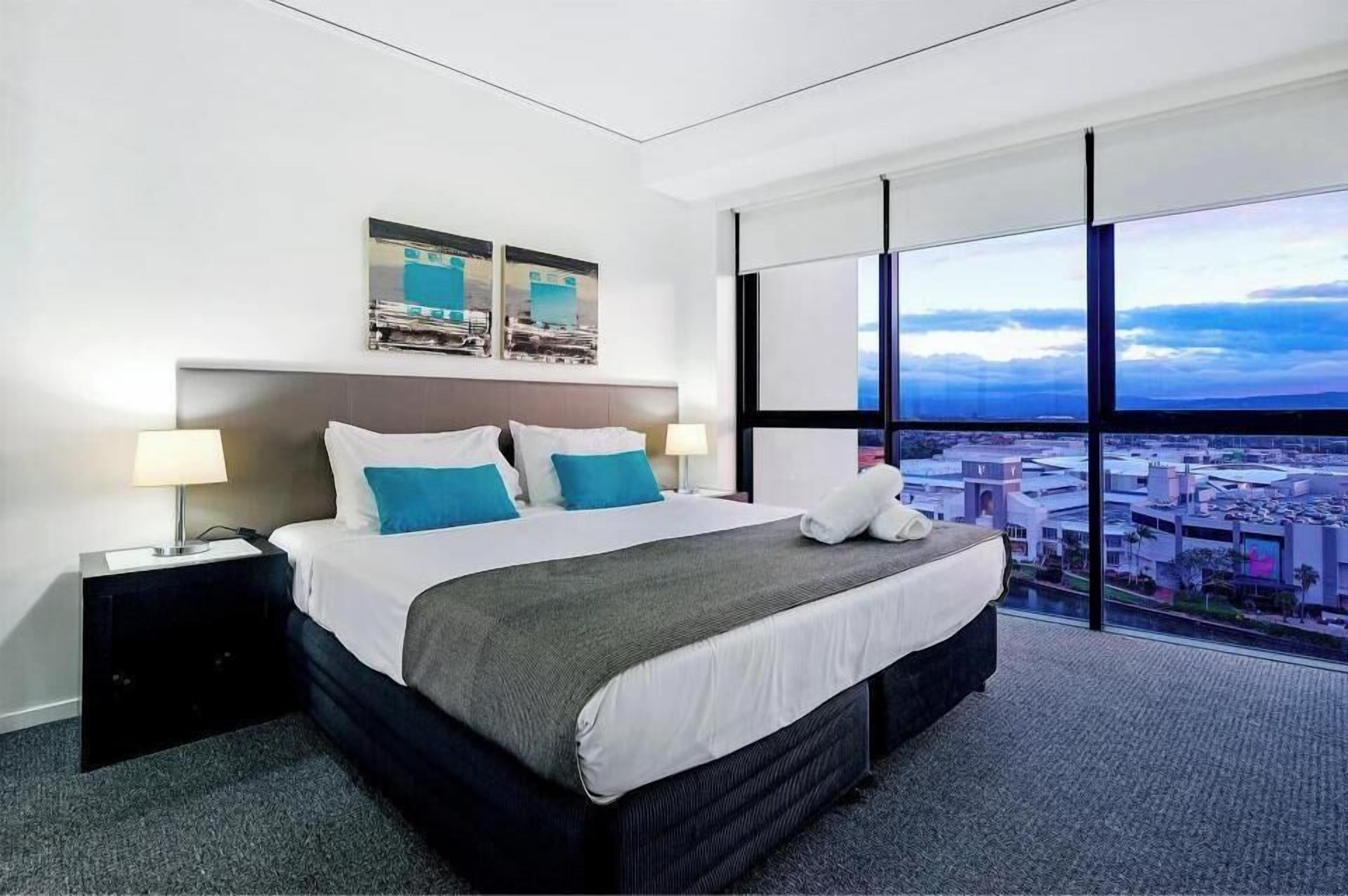 Ein modernes Hotelzimmer mit einem großen Bett, zwei Nachttischen, Blick auf die Stadt durch raumhohe Fenster und Kunstwerken über dem Bett.