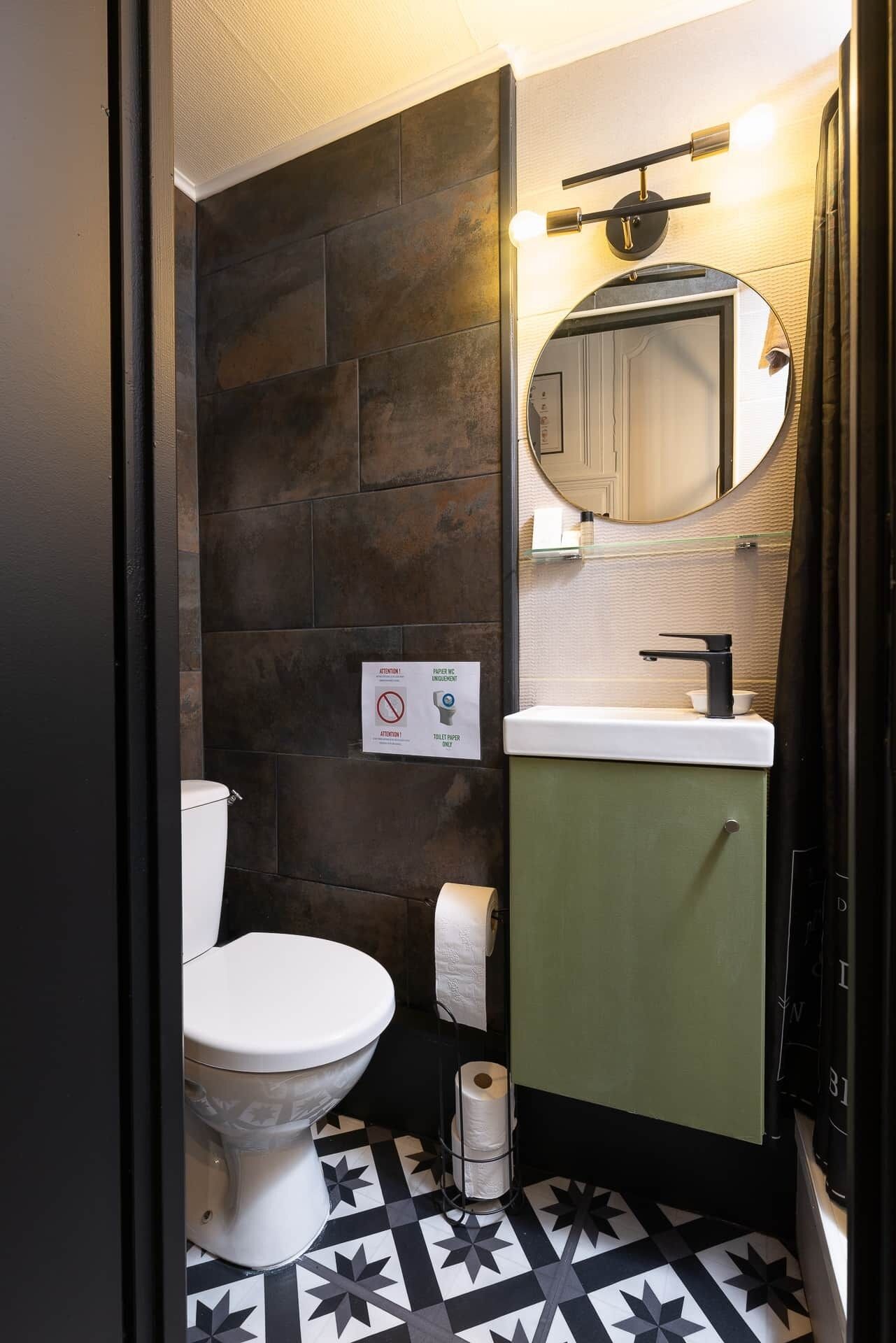 Une salle de bain avec des toilettes blanches, un meuble vert, un miroir rond et un sol à motifs noir et blanc.