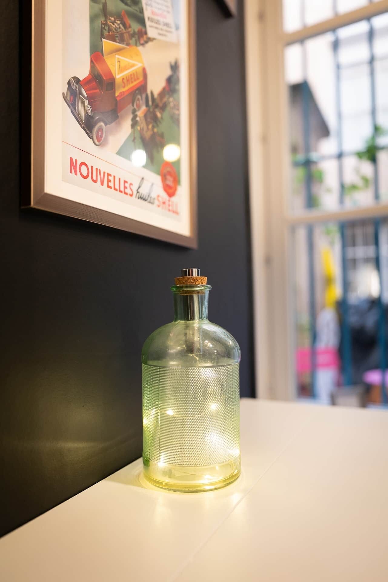 A framed advertisement for Shell on a wall, a clear glass bottle on a table, and a window with a view of buildings.