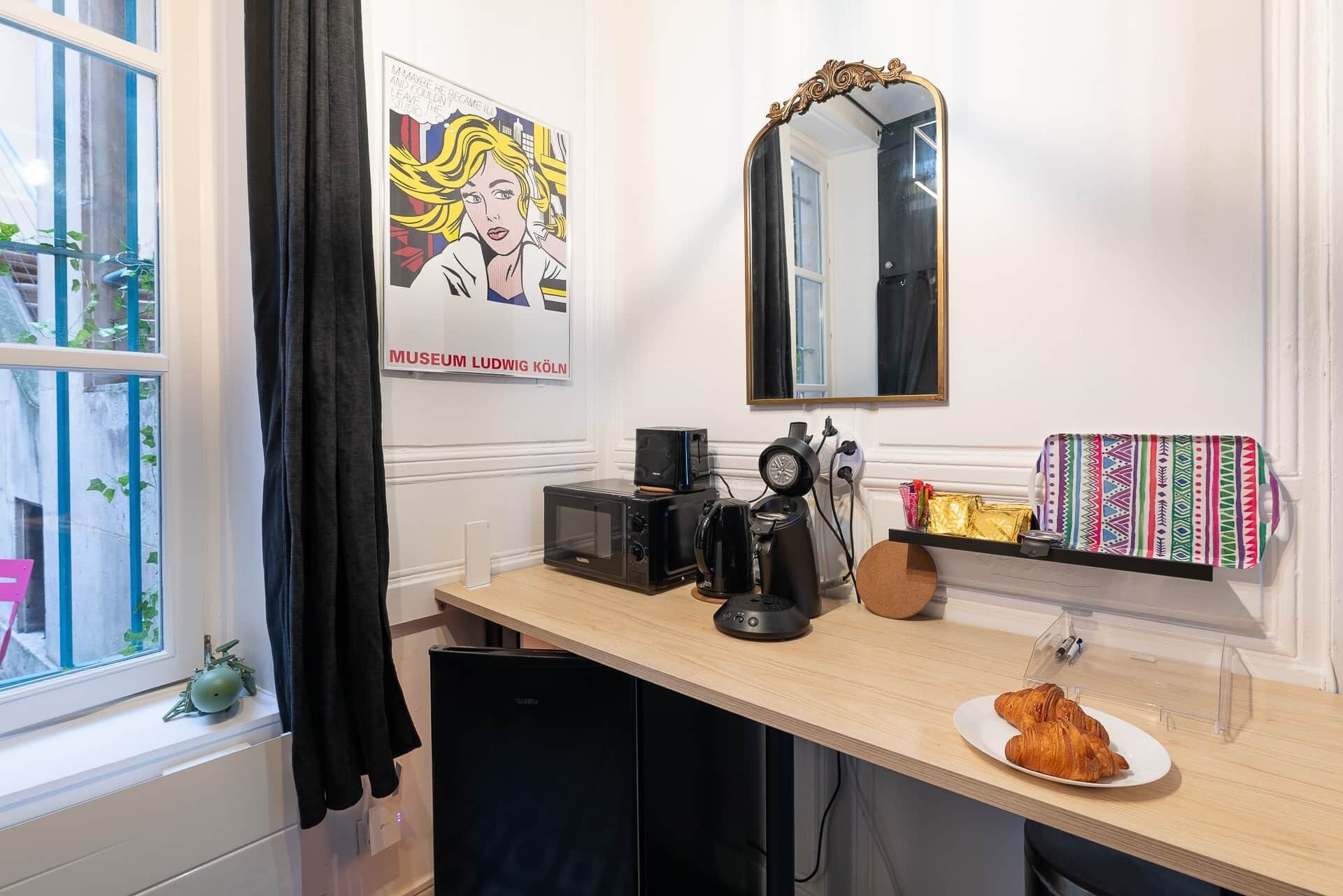 A compact hotel room with a kitchenette, a large mirror, a poster on the wall, and a window with curtains.
