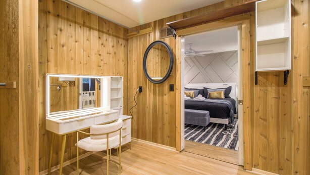 Une pièce aux murs en bois, une coiffeuse avec un miroir, un lit surmonté d’un tapis à rayures noires et blanches, et un ventilateur de plafond.