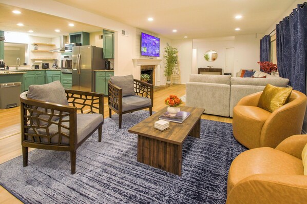 A modern living room with a sofa, armchairs, and a coffee table. A kitchen with green cabinets and a TV is visible in the background.