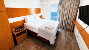 A hotel room with a bed, a TV, a wooden headboard, a nightstand, a suitcase, and a view of the ocean. - Hosts BR - Cobertura duplex frente mar (Fortaleza)