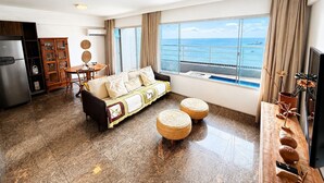 A living room with a sofa, dining table, and chairs, a kitchen area with a refrigerator, and a large window offering a view of the sea. - Hosts BR - Cobertura duplex frente mar (Fortaleza)