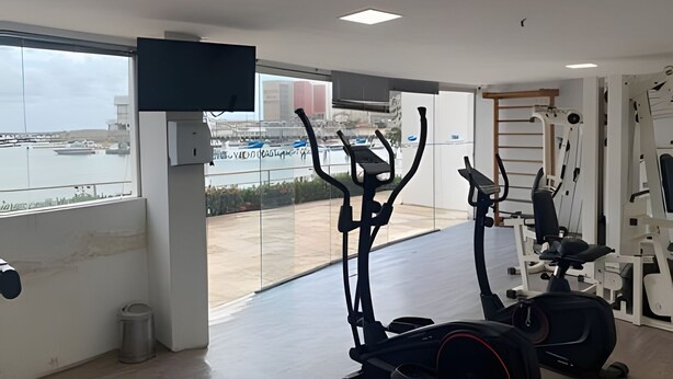 A gym with exercise equipment, a TV mounted on the wall, and a view of a marina through large windows. - Hosts BR - Flat Vista Mar no Meireles (Fortaleza)